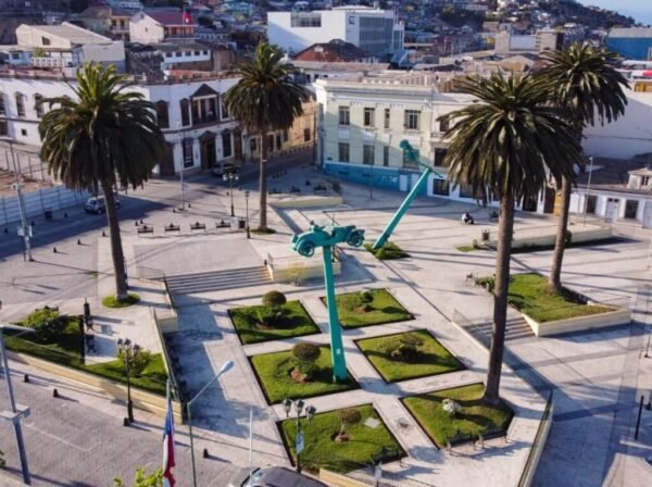 Vista aérea de una plaza en el Barrio Inglés de Coquimbo, destacando una moderna escultura de color turquesa en el centro rodeada de áreas verdes cuidadosamente diseñadas. Grandes palmeras flanquean la plaza, mientras los edificios históricos del barrio y la ciudad se extienden en el fondo bajo un cielo despejado.