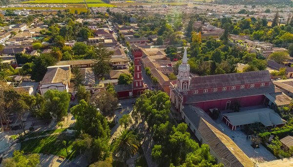 Vista aérea de la plaza de Vicuña, destacando la icónica iglesia roja con su torre y campanario, rodeada de árboles y un entorno urbano lleno de vegetación. En el fondo, se observan campos cultivados y casas que complementan la belleza del paisaje bajo una cálida luz del atardecer.
