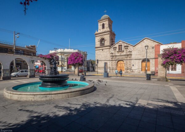 Vista de la Iglesia San Francisco en La Serena, destacada por su fachada de piedra y su torre campanario. En primer plano, una fuente con agua cristalina y detalles en bronce se complementa con árboles decorados con flores fucsias. La escena transcurre bajo un cielo despejado, reflejando la tranquilidad y el encanto del lugar.