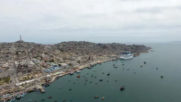 Vista aérea del puerto de Coquimbo, mostrando una bahía repleta de barcos pequeños y un crucero atracado en el muelle. Al fondo, se aprecia el icónico Monumento Cruz del Tercer Milenio en la cima de una colina, con la ciudad extendiéndose hacia el horizonte bajo un cielo parcialmente nublado.