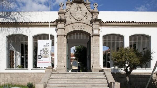 Fachada del Museo Arqueológico de La Serena, un edificio de estilo neocolonial con un imponente pórtico de piedra que lleva la inscripción 'Museo'. La entrada está enmarcada por ventanas con rejas y un jardín visible en su interior. El cielo azul despejado destaca la arquitectura histórica del lugar.