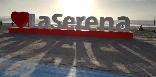 Escultura turística con el texto 'La Serena' en grandes letras blancas sobre una base roja, con un corazón rojo que reemplaza la palabra 'amo'. La instalación se encuentra frente a la playa, con el sol del atardecer brillando en el horizonte y proyectando sombras largas sobre el pavimento.