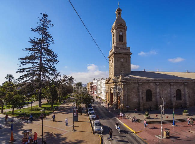 Vista de la Catedral de La Serena, Chile, capturada en un día soleado con cielo despejado. En la imagen se observa la histórica iglesia de piedra con su característica torre, rodeada de áreas peatonales, árboles y personas transitando.