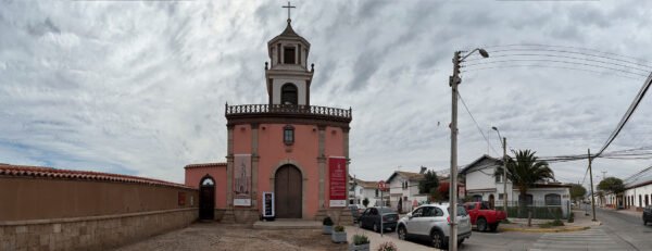 Vista panorámica de la Iglesia Santa Inés, un edificio histórico de estilo colonial con fachada rosada y una torre central coronada por una cruz. Ubicada en La Serena, está rodeada de calles tranquilas, vehículos estacionados y un cielo parcialmente nublado que resalta su belleza arquitectónica.