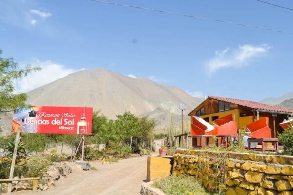 Exterior del restaurante solar 'Delicias del Sol' en Villaseca, rodeado de vegetación desértica y con paneles solares destacados en su terraza. En el fondo, se puede apreciar una imponente montaña bajo un cielo despejado, resaltando la conexión del lugar con la naturaleza y la energía renovable.