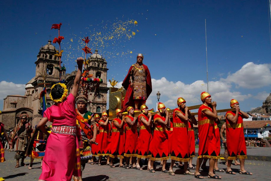 Representación del Inti Raymi, el festival del Sol Inca, con actores vestidos en trajes tradicionales rojos y dorados, llevando al Inca en un trono decorado. Al fondo, se observan las iglesias coloniales de Cusco bajo un cielo despejado, mientras pétalos de flores amarillas caen como parte de la ceremonia.