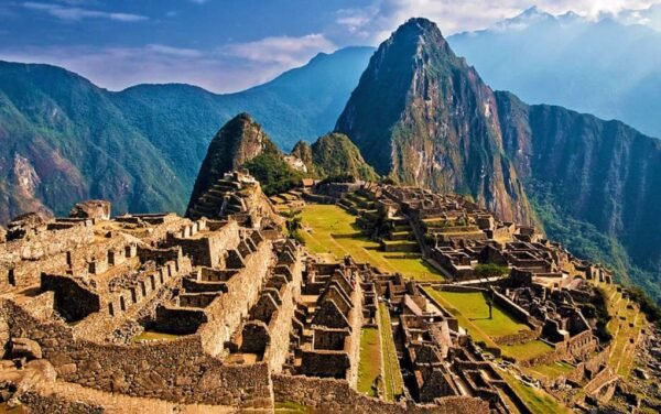 Vista panorámica de Machu Picchu, la antigua ciudadela inca enclavada en las montañas de los Andes peruanos. Las terrazas agrícolas, construcciones de piedra y senderos se destacan bajo la imponente presencia de la montaña Huayna Picchu, mientras la luz del sol ilumina el paisaje, resaltando su majestuosidad y conexión con la naturaleza.