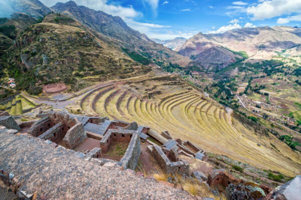 Vista panorámica de las terrazas agrícolas de Pisac, en el Valle Sagrado de los Incas, Perú. Las terrazas en forma de círculos y curvas se extienden por las laderas de las montañas, mientras el paisaje de valles fértiles y montañas majestuosas se despliega en el fondo bajo un cielo parcialmente nublado.